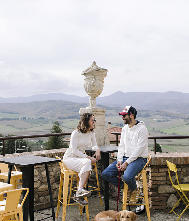 A couple sitting at an outdoor café in Lajatico, Tuscany, with panoramic rolling hills in the background and their dog resting at their feet.
