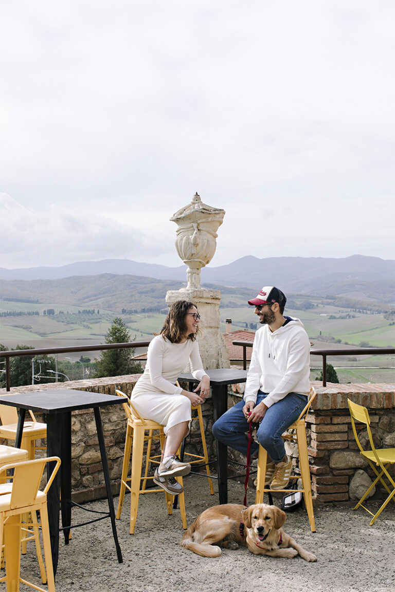A couple sitting at an outdoor café in Lajatico, Tuscany, with panoramic rolling hills in the background and their dog resting at their feet.